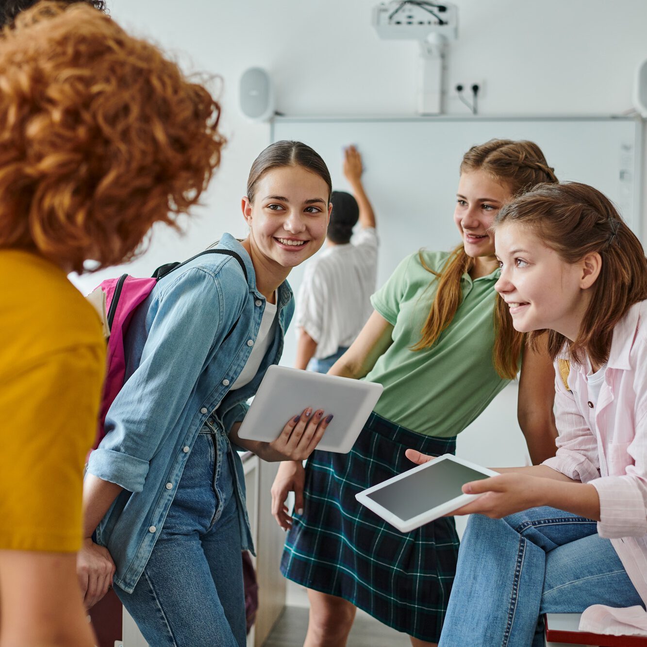 Smiling teen schoolgirls holding digital tablets near blurred friends in classroom in school